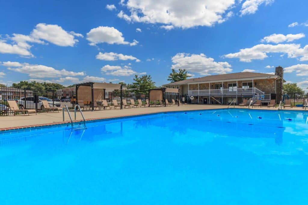 An outdoor swimming pool with clear blue water, surrounded by lounge chairs and a building in the background under a partly cloudy sky.