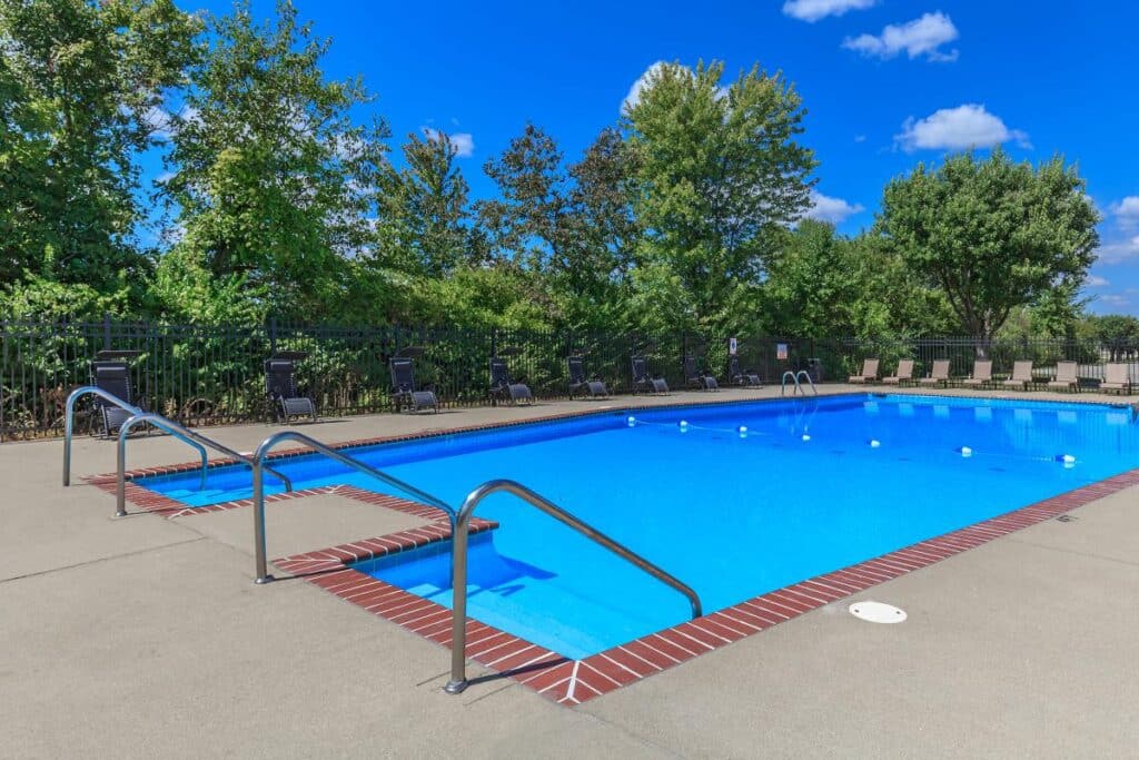 Rectangular outdoor swimming pool with metal handrails, lounge chairs along the perimeter, and trees in the background under a clear blue sky.