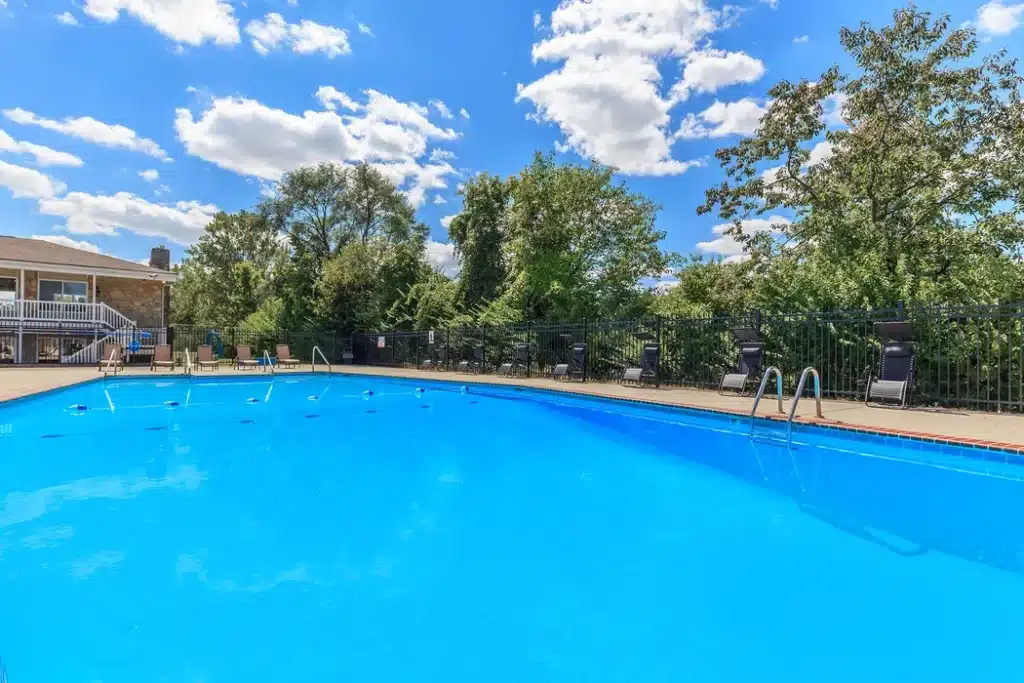 Outdoor swimming pool with clear blue water, surrounded by lounge chairs and trees under a mostly sunny sky with scattered clouds.