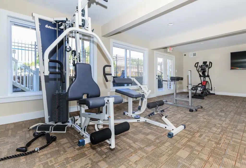 A small gym room with a multi-function weight machine, stationary bike, and elliptical trainer, brightened by large windows and neutral-colored walls.
