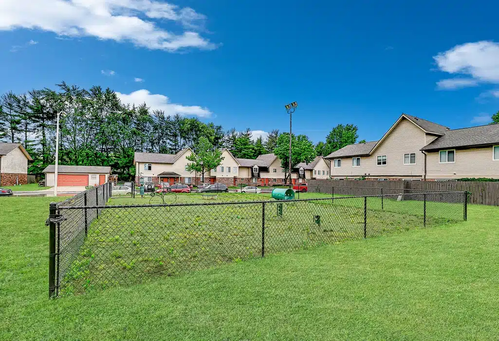 A small fenced-in area with a green waste bin sits in a grassy yard near residential houses under a blue sky with scattered clouds.