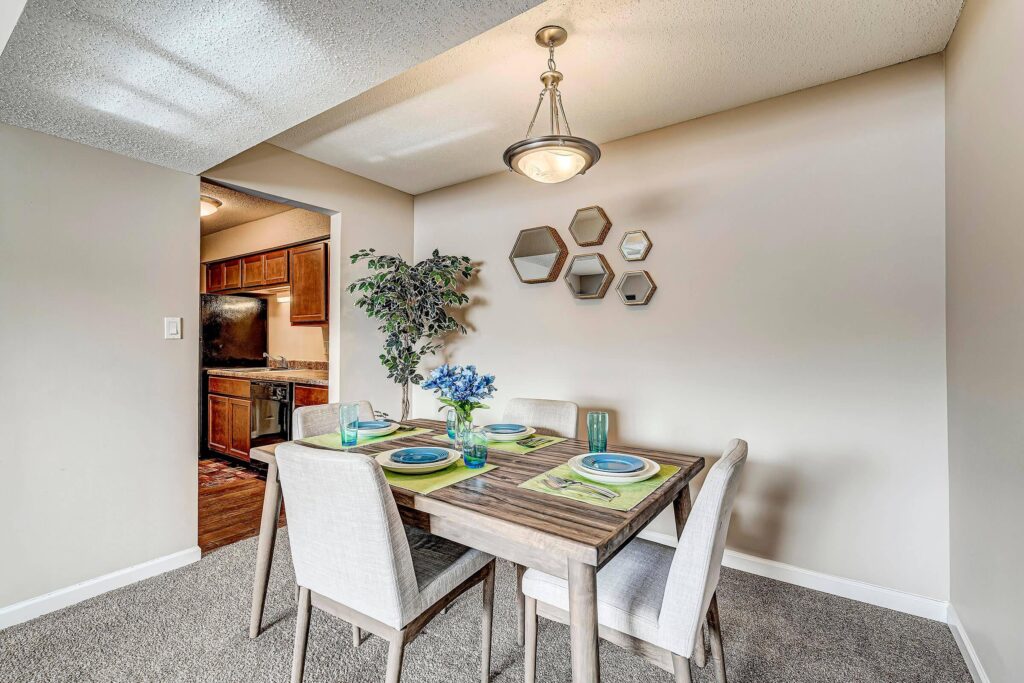 A small dining area with a wooden table set for four, beige chairs, a potted plant, and decorative hexagonal mirrors on the wall near an open kitchen.