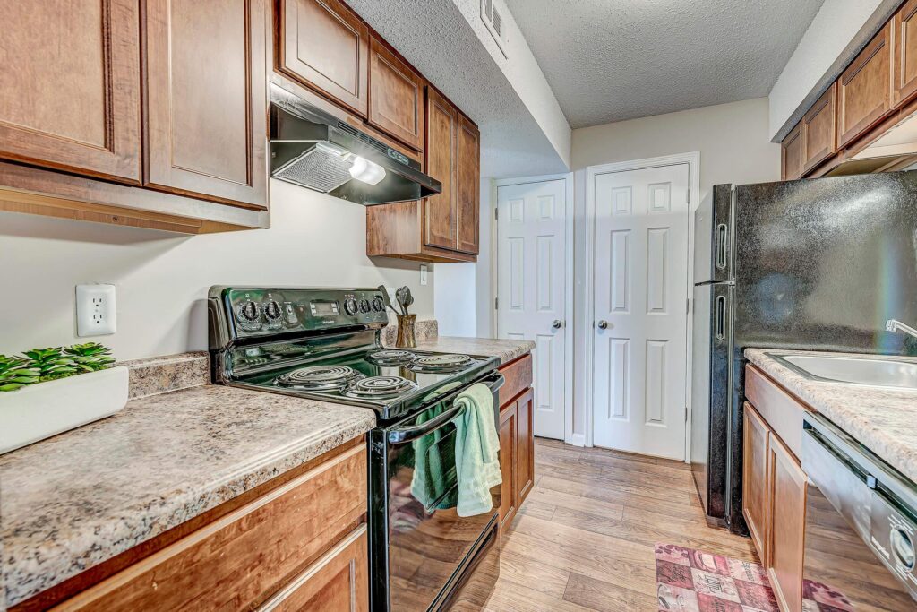 A galley kitchen with wood cabinets, black appliances, electric stove, double sink, marble-look countertops, and wood flooring. Two closed white doors are at the end of the kitchen.