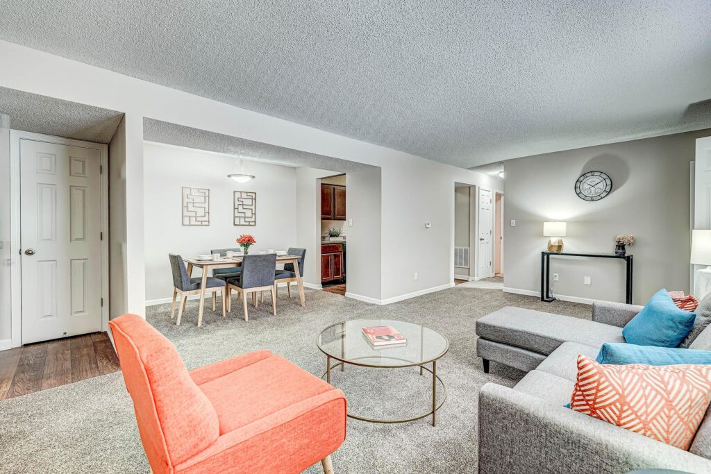 Modern living room with grey sofa, orange chair, glass coffee table, and adjacent dining area with four chairs; neutral walls and carpeted floor.