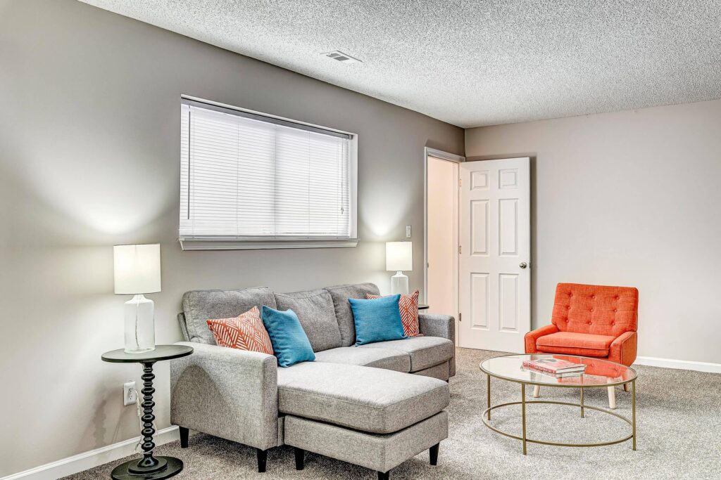 Living room with a gray sectional sofa, colorful accent pillows, orange armchair, round glass coffee table, two lamps, and a window with blinds.