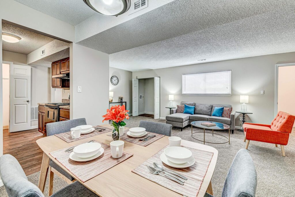 Modern apartment interior with a dining table set for four, an open-plan kitchen, and a living area with a gray sofa and orange chair.
