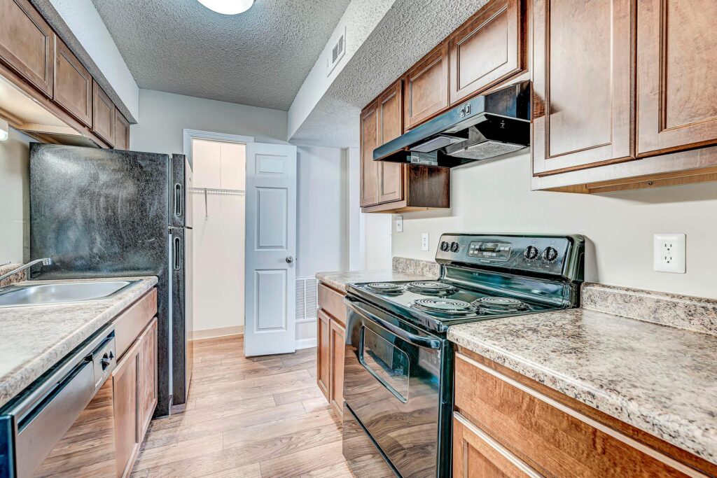 Galley kitchen with wood cabinets, black appliances, marble-look countertops, and wood flooring. Open door reveals pantry with shelving in the background.