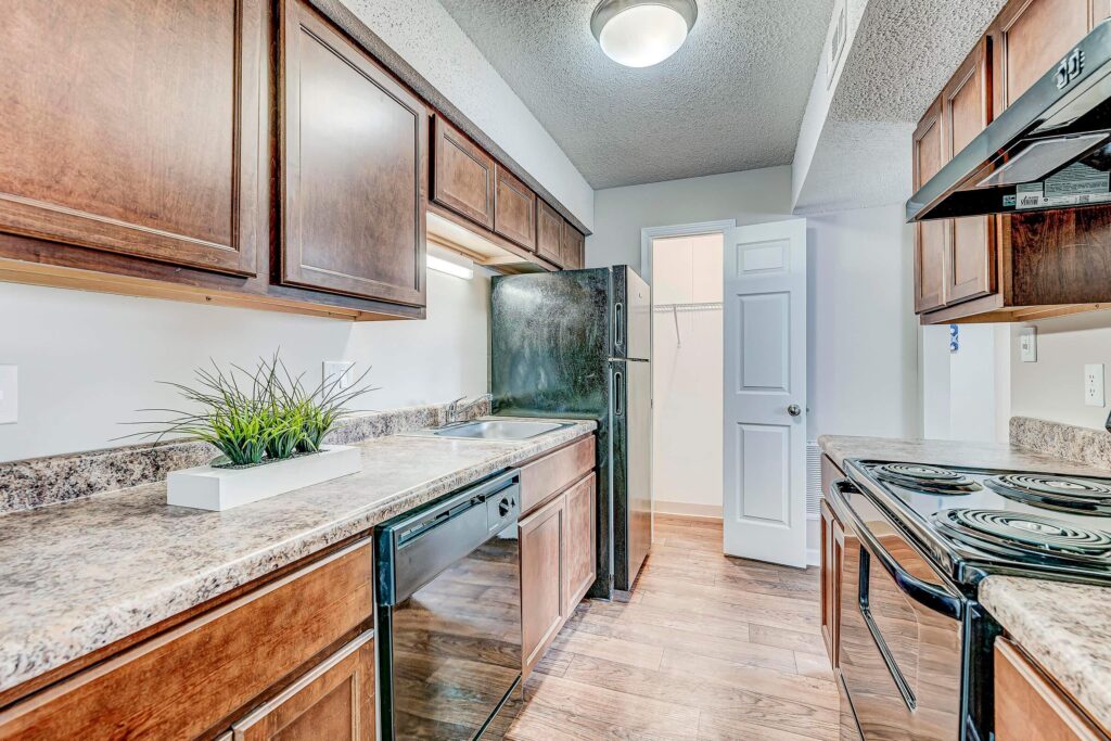 Galley-style kitchen with dark wood cabinets, black appliances, marble-patterned countertops, and a potted plant next to the sink; open pantry door in the background.