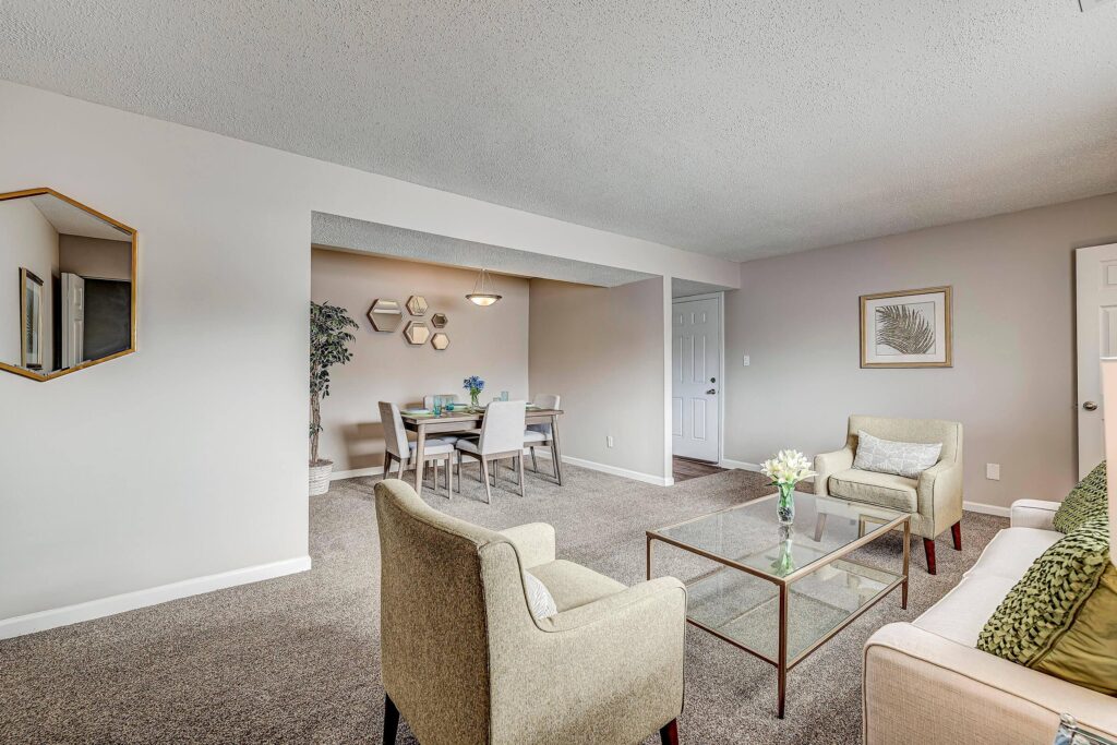 A living room with beige carpet, two chairs, a sofa, a glass coffee table, and a dining area with a table and four chairs in the background.