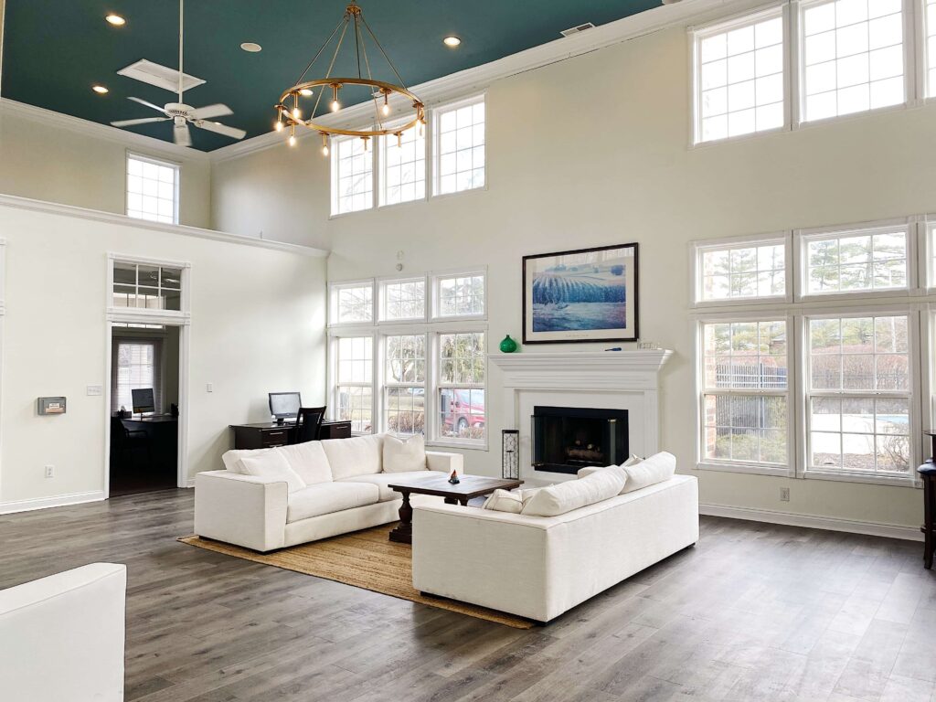 Spacious, well-lit living room with high ceilings, large windows, white sofas, a fireplace, wall art, and wood flooring. A desk is in the corner by the window.