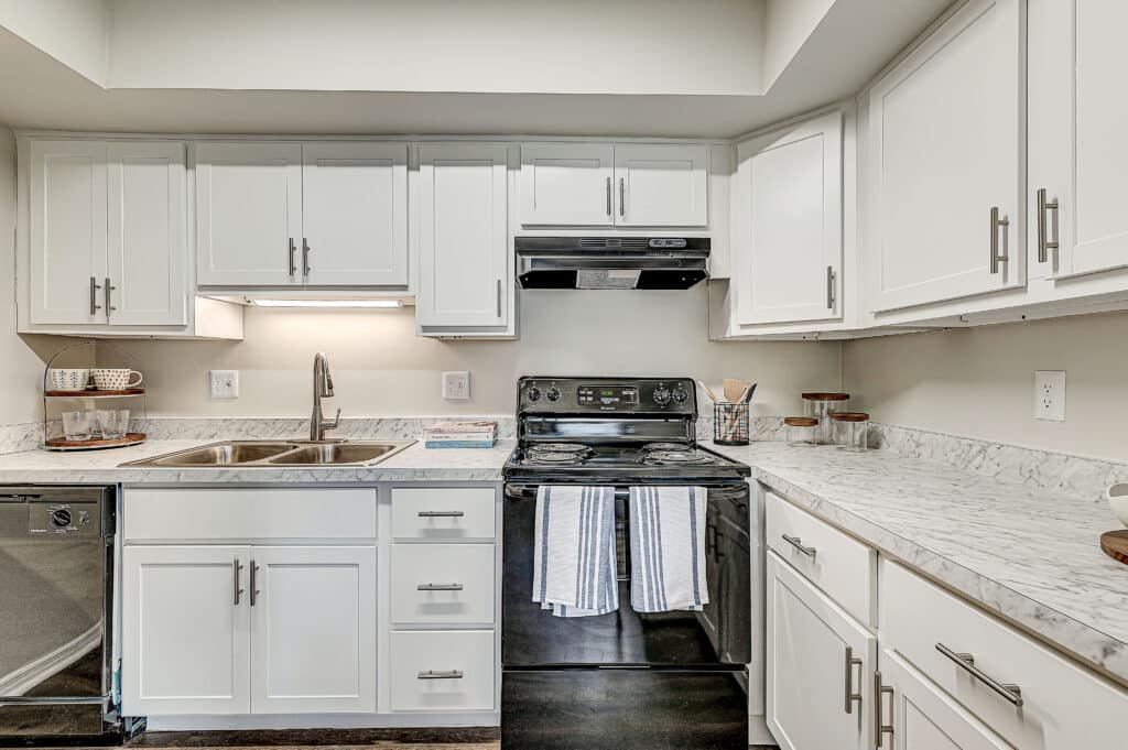 Modern kitchen at Stonehedge Apartments featuring white cabinets, marble countertops, stainless steel sink, black stove and range hood, with various utensils neatly arranged on the counters.