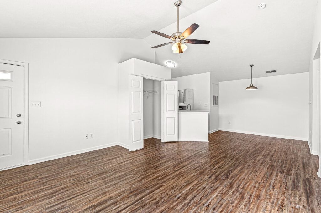 Unfurnished room with wood-style flooring, white walls, ceiling fan with lights, open closet, and a small kitchen area visible in the background.