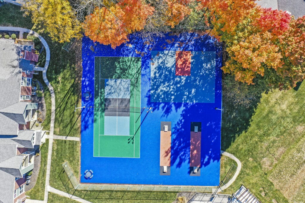 Aerial view of a blue sports court with tennis and basketball areas at Enclave at Eagle Creek, surrounded by autumn trees and nearby residential buildings.