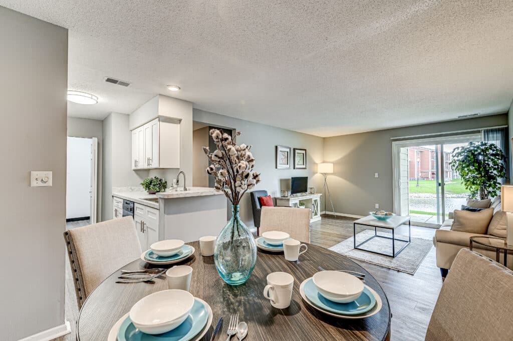 Dining table set for four in a modern open-plan living space at Villages of Bent Tree Apartments, featuring a kitchen, living area, and sliding glass doors leading to a patio.