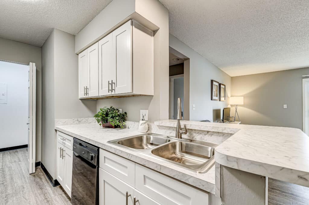 Modern kitchen at Villages of Bent Tree Apartments with white cabinets, marble countertops, double stainless steel sink, built-in dishwasher, and potted plant next to an open living area.