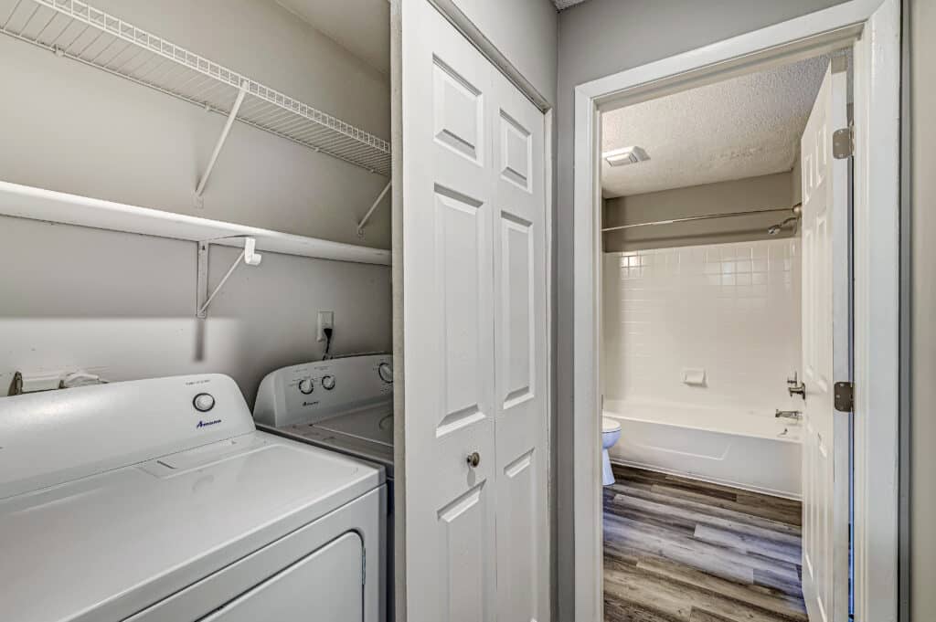 Laundry area with a washing machine and dryer next to an open door leading to a bathroom with a bathtub and white tile walls at Villages of Bent Tree Apartments.