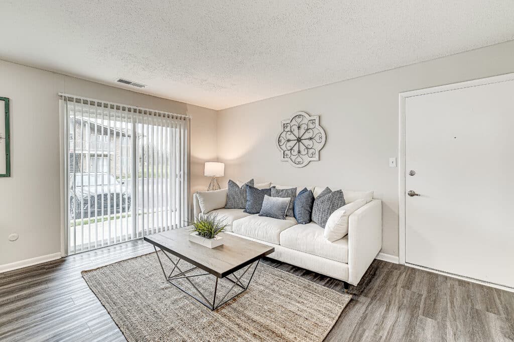A modern living room at Ninth Avenue Apartments features a white sofa, gray pillows, a wooden coffee table, a floor lamp, wall art, and a large window with vertical blinds.