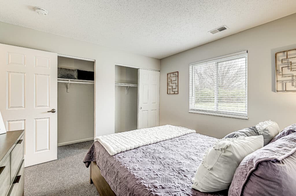 Bedroom at Ninth Avenue Apartments with a neatly made bed, two open closets, a window with blinds, and minimal decor on cream-colored walls.