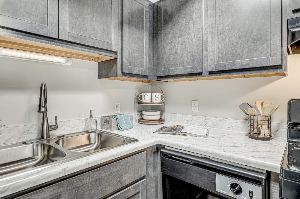 Modern kitchen at Ninth Avenue Apartments featuring gray cabinets, a marble countertop, stainless steel sink, dish rack, utensil holder, and an open cookbook on the counter.