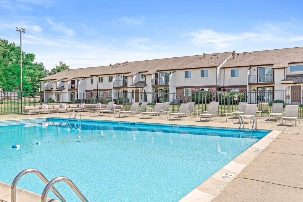 Outdoor swimming pool with clear blue water, surrounded by lounge chairs and a fence, with a row of two-story townhouses in the background under a blue sky.