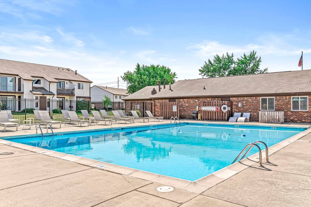 Outdoor swimming pool with lounge chairs on a concrete deck, surrounded by apartment buildings and a brick pool house under a clear blue sky.