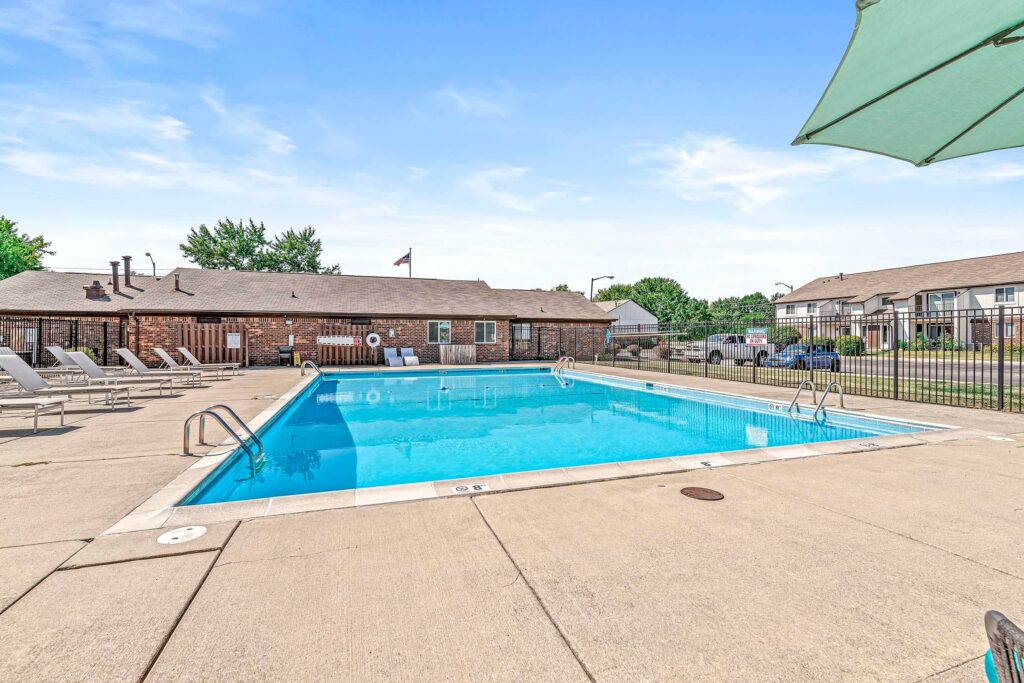 Outdoor swimming pool with clear water, surrounded by lounge chairs, a fence, and apartment buildings in the background under a blue sky.