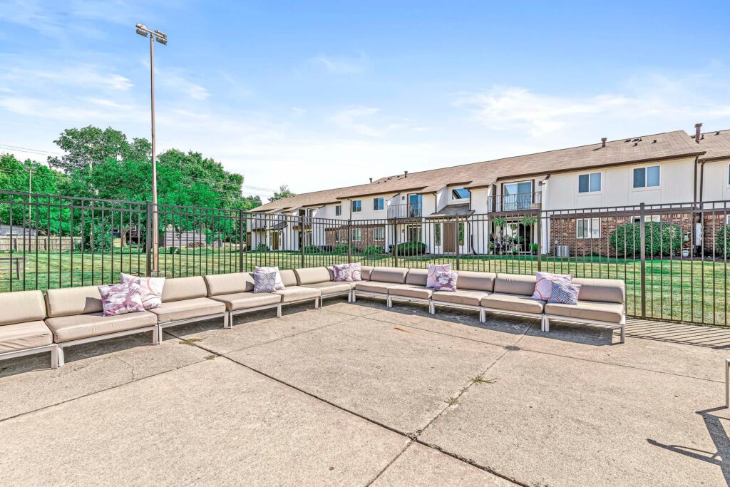 Outdoor seating area with beige cushioned benches arranged in an L-shape, accented with decorative pillows, enclosed by a metal fence, with apartment buildings and grass in the background.