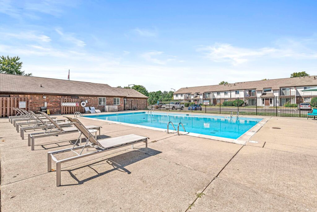 Outdoor swimming pool with a concrete deck, several lounge chairs, and nearby residential buildings on a clear day.