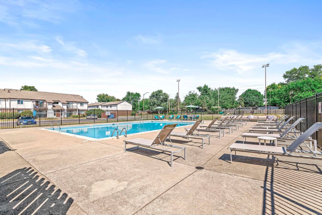 Outdoor swimming pool with lounge chairs on a concrete deck, enclosed by a black metal fence, with trees and buildings in the background under a clear sky.