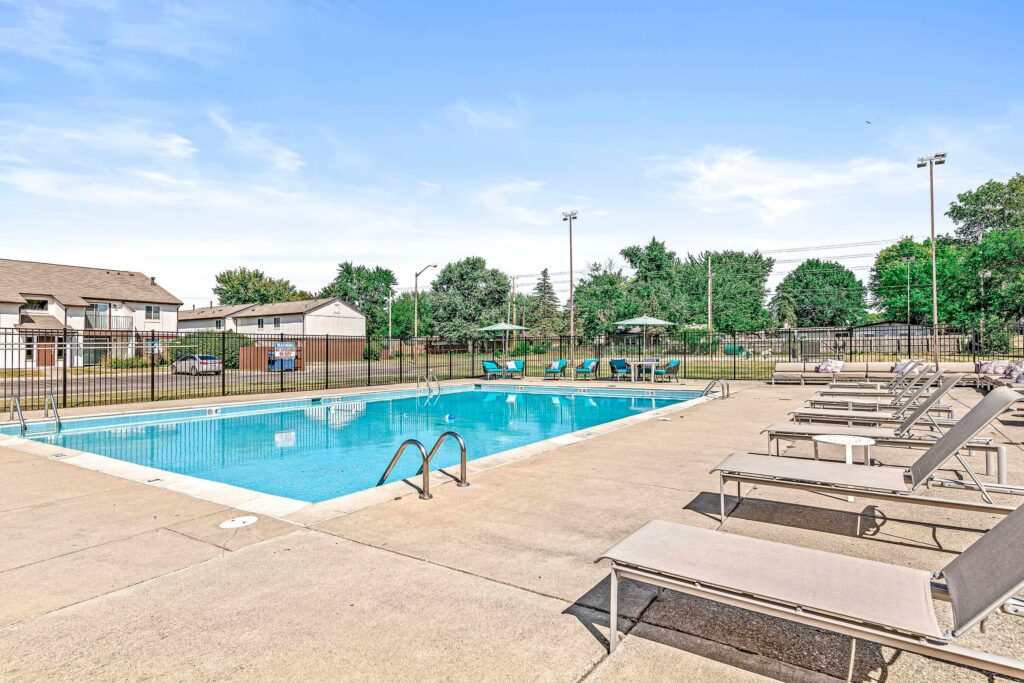 Outdoor swimming pool with metal ladders, surrounded by lounge chairs and umbrellas, bordered by a fence, with trees and residential buildings in the background under a clear sky.
