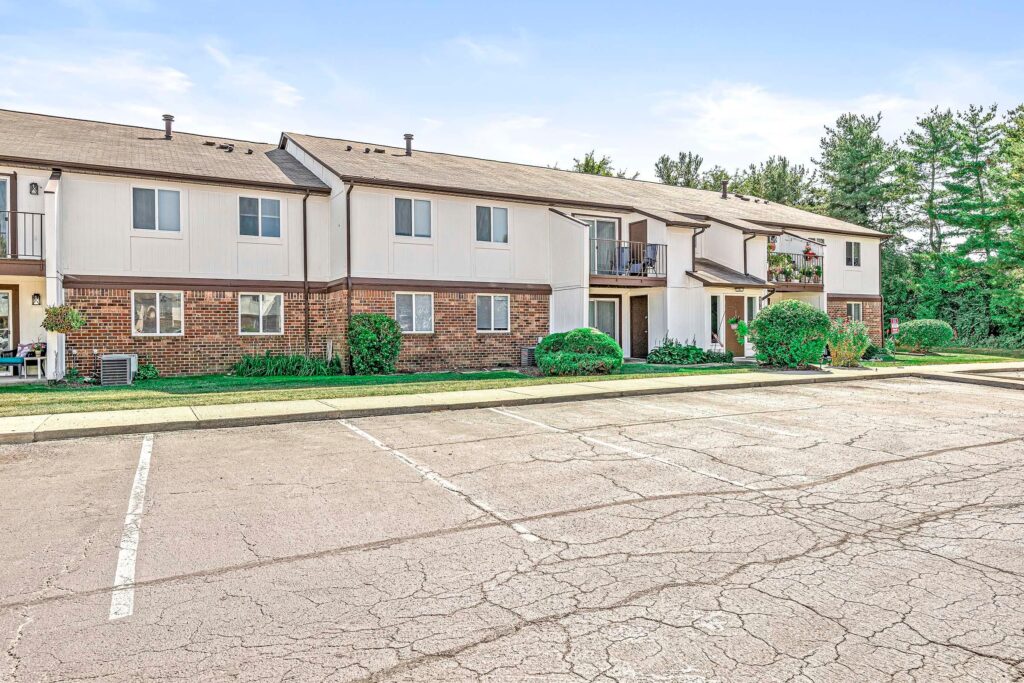 Two-story apartment building with brick and siding exterior, small balconies, landscaped bushes, and an empty parking lot in front.