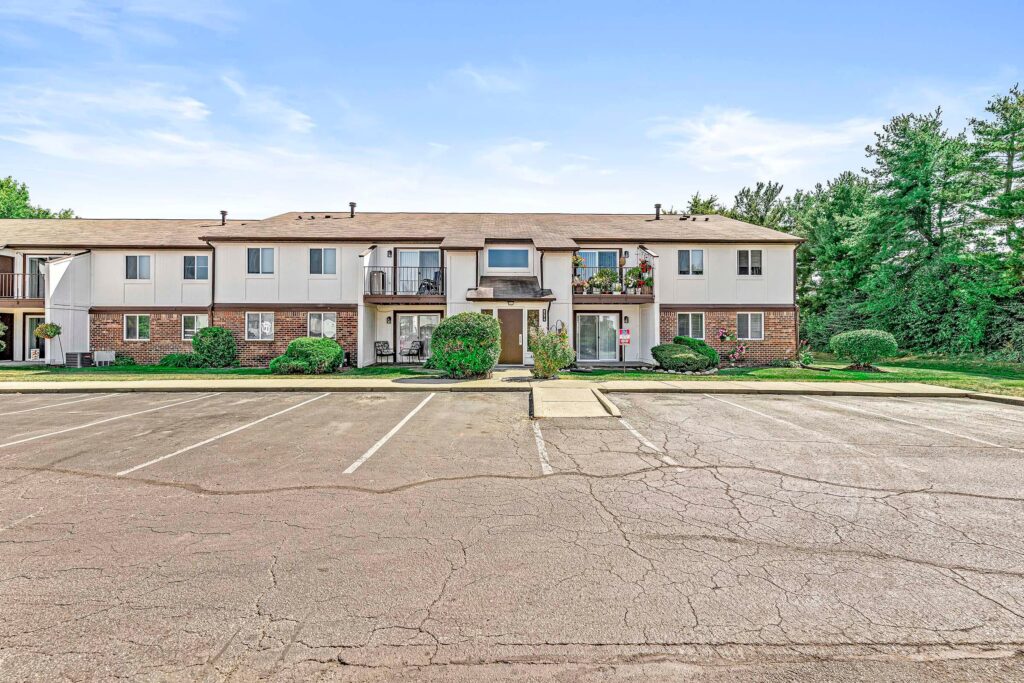 Two-story apartment building with brick and white siding exterior, balconies, and a mostly empty parking lot in front, surrounded by trees.
