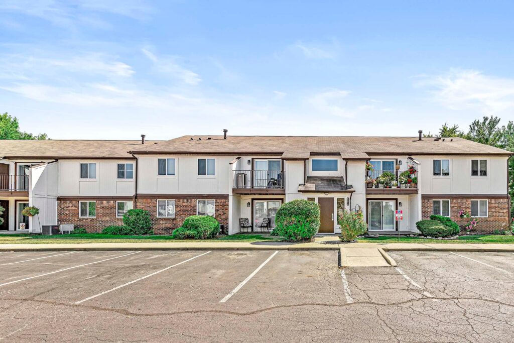 Two-story apartment building with brick and white siding exterior, balconies, shrubs, and an adjacent parking lot with empty spaces under a clear sky.