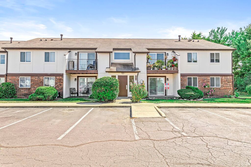 Two-story apartment building with brick and white siding exterior, balconies, landscaped bushes, and an empty parking lot in front.