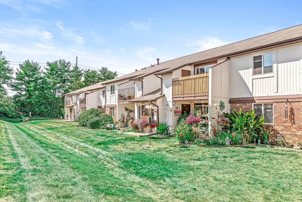 Row of two-story townhouse units with balconies and patios, surrounded by a grassy lawn and landscaped gardens, on a sunny day.