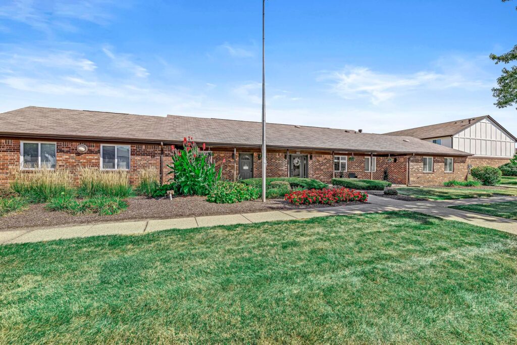 Single-story brick building with a brown roof, surrounded by landscaped gardens, grass, and a concrete walkway under a blue sky.