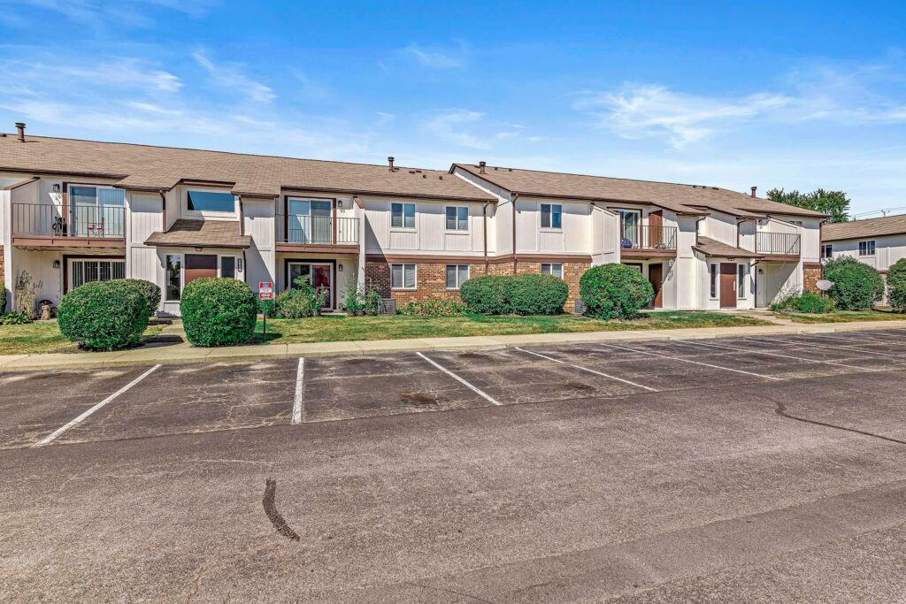 A row of two-story townhouses with balconies, trimmed bushes in front, and an empty parking lot under a clear blue sky.