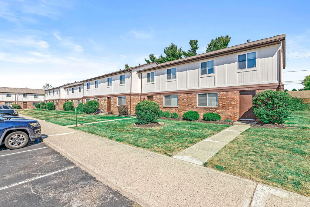 A row of two-story brick and white townhouses with small shrubs and grass lawns, adjacent to a parking lot under a clear sky.