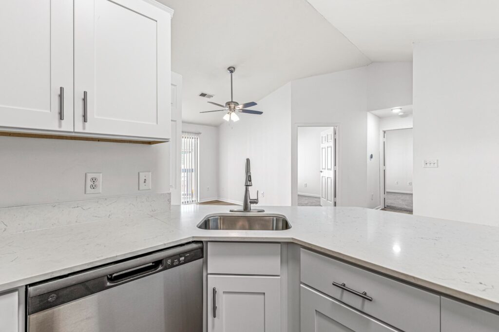Modern kitchen with white cabinets, stainless steel dishwasher, undermount sink, and quartz countertops, overlooking an open living space with ceiling fan and visible doorways.