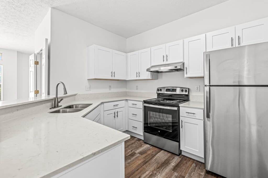 Modern kitchen with white cabinets, stainless steel appliances including a stove and refrigerator, a double sink, and light-colored countertops with wood-look flooring.