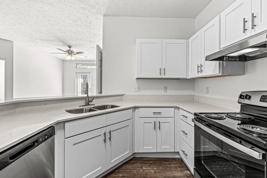 Modern kitchen with white cabinets, stainless steel appliances, electric stove, and a double sink beneath a pass-through window. Dark wood flooring contrasts with the light countertops.