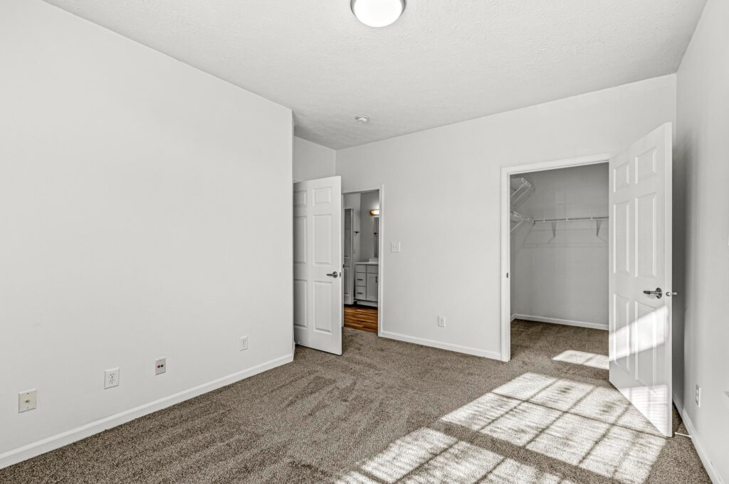 Empty, carpeted bedroom with white walls, two doors—one open to a walk-in closet, the other to a hallway—and sunlight streaming in from an unseen window.