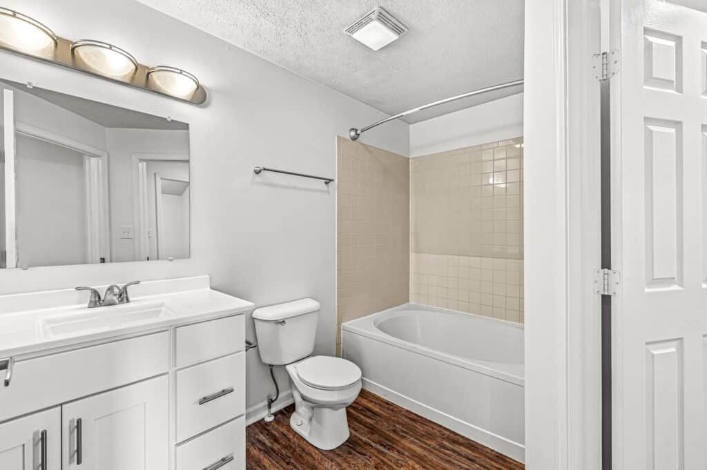 A bathroom with a white vanity, toilet, and bathtub with beige tile surround; wood-style flooring and overhead lighting are visible.