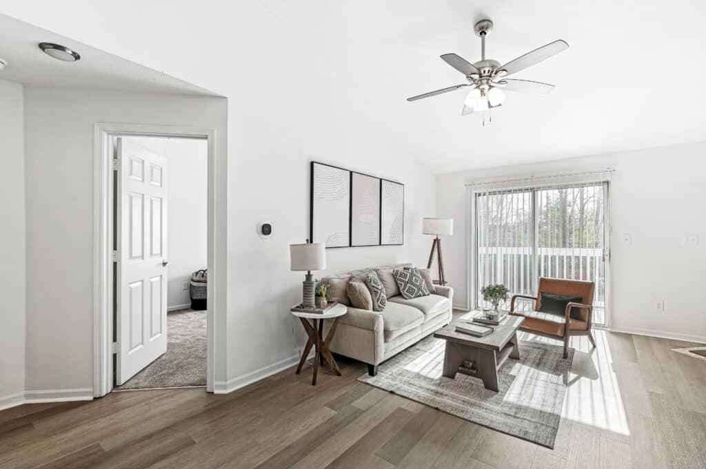 Modern living room at Buffalo Creek Apartments with a sofa, armchair, coffee table, and lamps. Features large windows with blinds, wood flooring, ceiling fan, and an adjacent room visible through an open door.