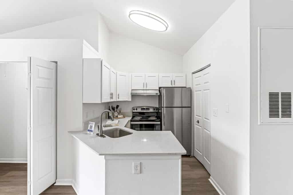 Modern kitchen at Buffalo Creek Apartments features white cabinets, stainless steel appliances, quartz countertops, and a double sink under a circular ceiling light. Closet and utility panel are conveniently located on the right.