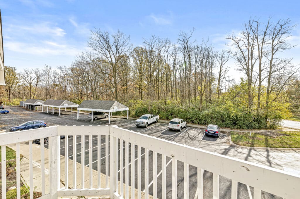 View from a white balcony at Buffalo Creek Apartments, overlooking a parking lot with several cars, carports, and trees in the background under a clear blue sky.