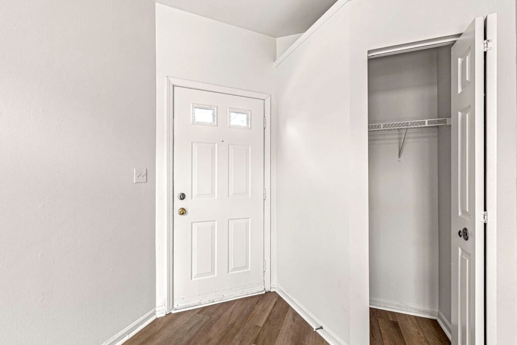 A white front door with three square windows is next to an open closet with a white door and a wire shelf. The floor is wood with a light brown finish.