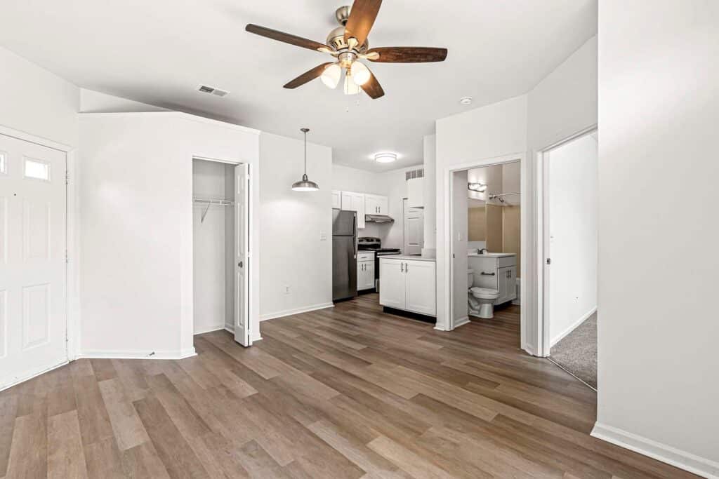 Open-plan apartment with wood-style flooring, ceiling fan, kitchen area with stainless steel appliances, two open doorways, a closet, and a view into a bathroom.