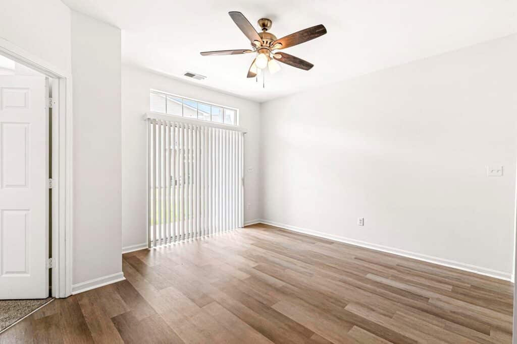 Empty room with white walls, wood-look flooring, ceiling fan with lights, window with vertical blinds, and a door leading outside.