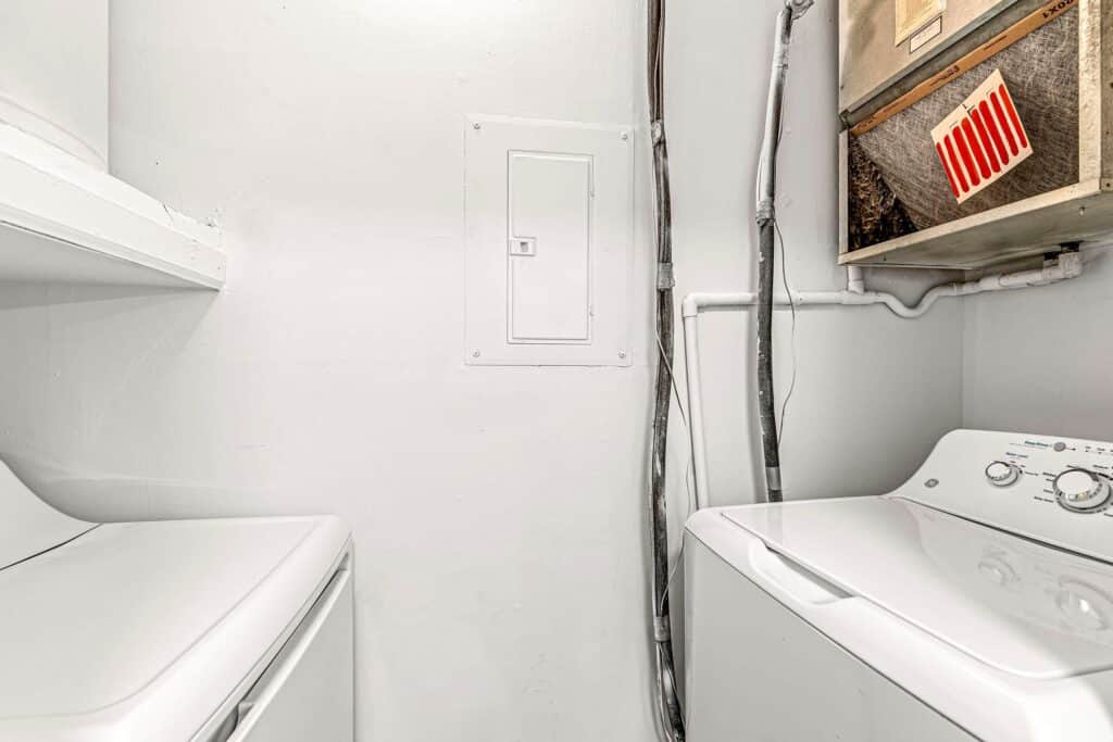 A small laundry room with a white washing machine and dryer, exposed pipes, an electrical panel, and a furnace with a red filter on the upper right.